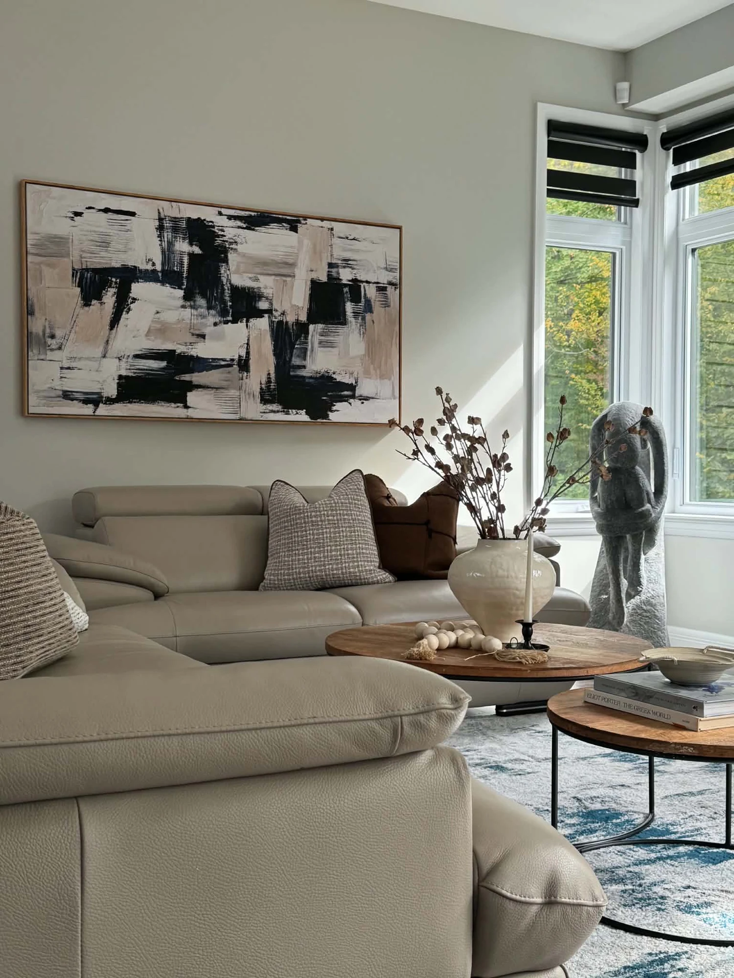 Living room with a cream sectional, round wood tables and abstract artwork beside tall windows